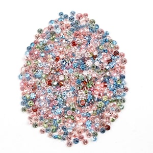 May include: A collection of small, faceted gemstones in various colors, including pink, blue, green, and red, are scattered on a white surface. The gemstones are round and appear to be cut with multiple facets, creating a sparkling effect. The overall arrangement is dense and visually appealing.