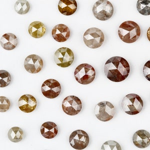 May include: Assortment of round, faceted, rough diamonds in various shades of brown, grey, and yellow. The diamonds are scattered on a white surface.