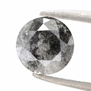 May include: A round, salt and pepper diamond held by silver tweezers. The diamond has a faceted cut with a mix of gray and white tones. The gem is set against a white background.