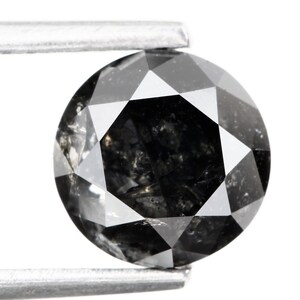 May include: A round, faceted black diamond held by silver tweezers against a white background. The gemstone has a complex, multi-faceted surface that reflects light, showcasing its dark color and internal inclusions.
