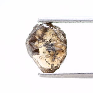 May include: A brown, faceted, rough diamond held by tweezers against a white background.