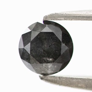 May include: A black round diamond with a faceted cut, held by tweezers against a white background.