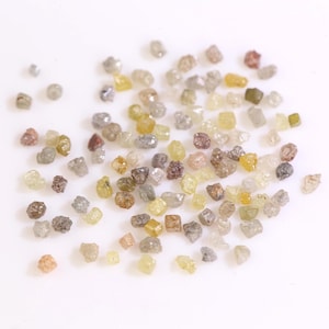 Puede incluir: Una colección de diamantes en bruto sin cortar en varios colores, incluyendo amarillo, marrón, gris y transparente. Las piedras preciosas están dispersas sobre una superficie blanca, mostrando sus formas y texturas naturales e irregulares. Estos diamantes en bruto son adecuados para la fabricación de joyas.