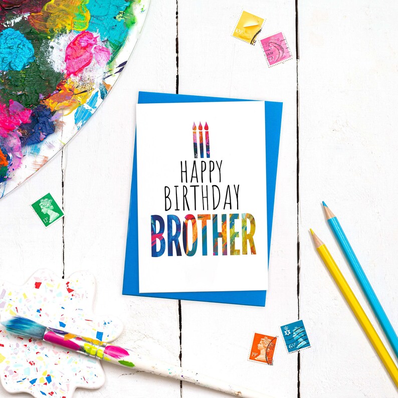 May include: A white card with a blue border and the text "Happy Birthday Brother" in black lettering. The text is surrounded by colorful abstract shapes. Three colorful candles are depicted above the text.
