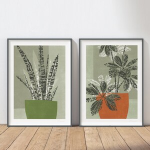 May include: Two framed prints of botanical illustrations. The print on the left features a green and black plant in a green pot. The print on the right features a green and white plant in an orange pot.