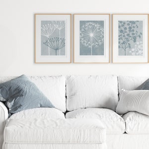 May include: Three framed prints with a light blue background and white floral designs. The prints are hanging on a white wall above a white couch with blue throw pillows.