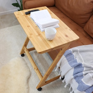 May include: A light brown wooden side table with wheels, a book, a white mug, and a remote control on top. The table is next to a brown leather sofa with a white and blue striped throw blanket.