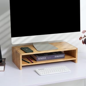 May include: A wooden monitor stand with two shelves. The top shelf holds a smartphone and a computer monitor. The bottom shelf holds a book titled "Rebecca" by Daphne du Maurier and a few pens. A white keyboard is on the desk below the stand.