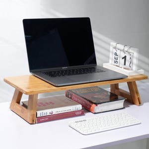 May include: A bamboo laptop stand with a laptop, books, and a white keyboard on a white desk. The stand has a shelf for books and a raised platform for a laptop. The calendar on the stand reads "SEP 12".