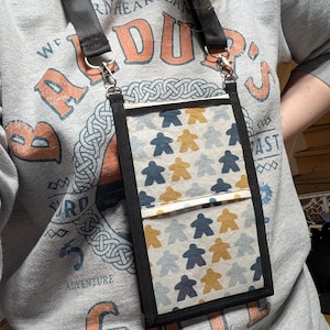 Könnte beinhalten: Eine schwarze Umhängetasche für Telefone mit einer gemusterten Vorderseite mit blauen und goldenen Spielfiguren auf hellem Hintergrund. Die Tasche hat einen schwarzen Riemen und Besatz sowie eine kleine Tasche. Die Person trägt ein graues Sweatshirt mit orangefarbenem und blauem Text.