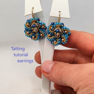 May include: A pair of blue and gold beaded earrings with a floral design. The earrings are hanging from a white display stand.