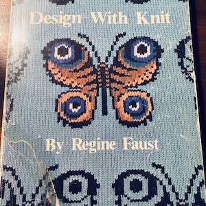 May include: A blue book with a knitted butterfly design on the cover. The title is "Design With Knit" by "Regine Faust".