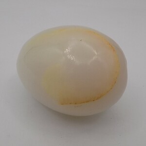 May include: A single, oval-shaped, hard-boiled egg with a smooth, off-white shell. The egg has a slight yellow discoloration on one side, likely from the cooking process. The egg is set against a plain white background.