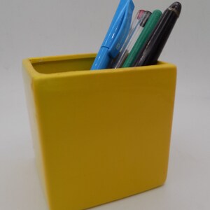 Può includere: Un portapenne in ceramica giallo con quattro penne al suo interno. Le penne sono blu, rosse, verdi e nere.