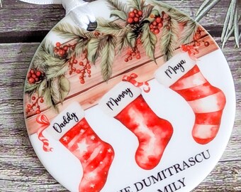 May include: A round Christmas ornament featuring three red and white stockings labeled "Daddy," "Mummy," and "Maya." The ornament has a festive design with greenery and berries, and the text "THE DUMITRASCU FAMILY."