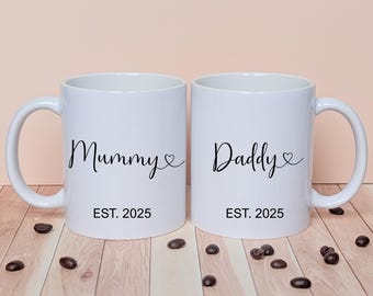 May include: Two white ceramic mugs with black script that reads "Mummy" and "Daddy" with a heart symbol. The mugs also have "EST. 2025" printed below the names. Coffee beans are scattered around the mugs.
