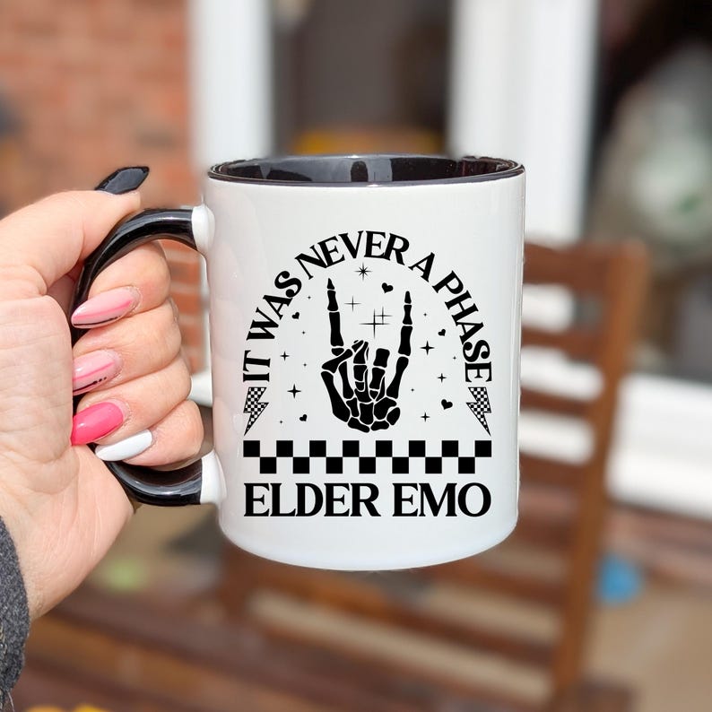 May include: White ceramic mug with a black interior and handle. The mug features the text "IT WAS NEVER A PHASE ELDER EMO" with a skeleton hand making a rock and roll hand gesture. The design includes stars and a checkered pattern.