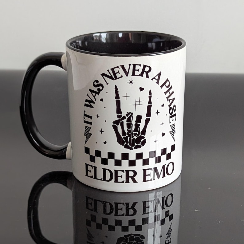 White ceramic mug with a black handle and interior. The mug features the text 'IT WAS NEVER A PHASE ELDER EMO' in black, along with a skeleton hand rock and roll symbol, lightning bolts, and a checkerboard pattern.