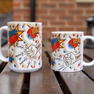 May include: Two white ceramic mugs with the words "SUPER DAD!" in a comic book style. The larger mug has names like Paulina and Martyna. The smaller mug has names like Sophia and Zakariya. Both mugs have a "SPECIAL EDITION" banner.