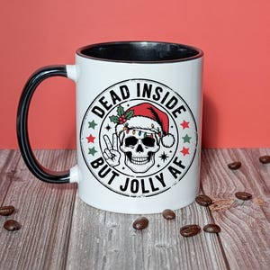 May include: White ceramic mug with a black handle and rim. The mug features a graphic of a skull wearing a Santa hat, with the text "DEAD INSIDE BUT JOLLY AF" in a circular design. The design includes stars and holly.