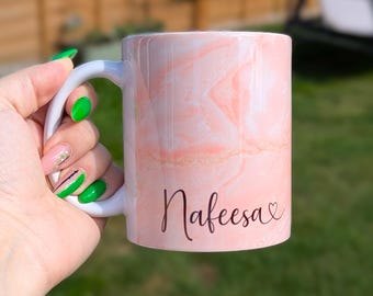 May include: A white ceramic mug with a pink marble design and the name "Nafeesa" in a cursive font. The mug has a white handle and is held by a hand with green and pink nail polish.