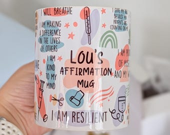 May include: White ceramic mug with colorful illustrations and text. The mug features affirmations, a thermometer, a rainbow, and the text "Lou's Affirmation Mug". The mug is held by a person.