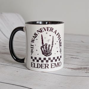 May include: White ceramic mug with a black handle and rim. The mug features the text "IT WAS NEVER A PHASE ELDER EMO" in black, along with a black hand gesture and decorative elements. The mug has a checkered pattern at the bottom.