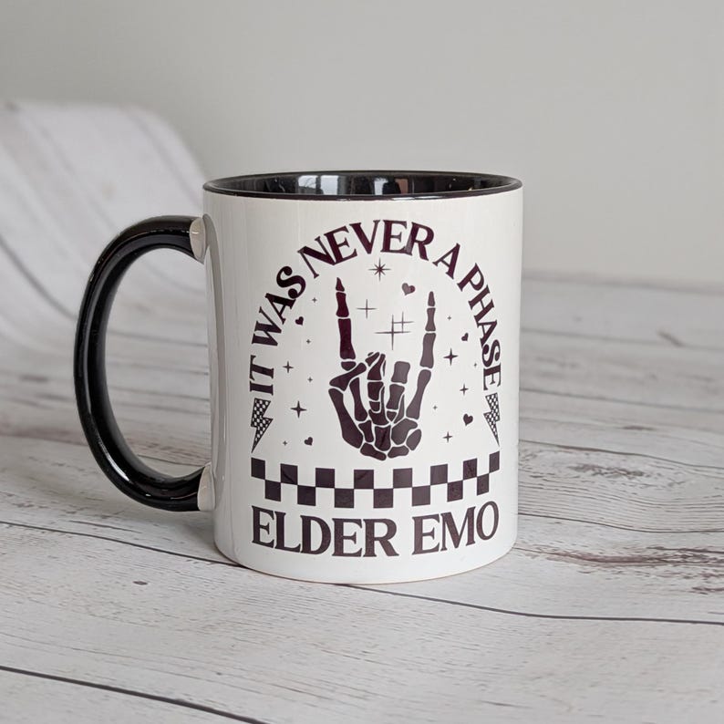 May include: White ceramic mug with a black handle and rim. The mug features the text "IT WAS NEVER A PHASE ELDER EMO" in black, along with a black hand gesture and decorative elements. The mug has a checkered pattern at the bottom.