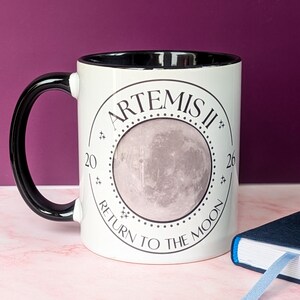 May include: White ceramic mug with a black handle and rim. The mug features the text "ARTEMIS II" and "RETURN TO THE MOON" around a gray image of the moon. The numbers "20" and "26" are also printed on the mug.