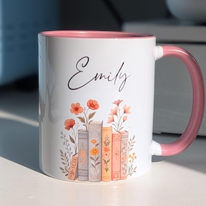 May include: White ceramic mug with a pink interior and handle. The mug features the name "Emily" in cursive script. A floral design of books and flowers decorates the front of the mug, with orange and pink flowers.