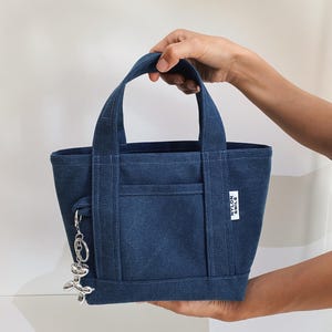 K&ouml;nnte beinhalten: Eine dunkelblaue Tragetasche aus Denim mit zwei stabilen Griffen und einer Au&szlig;entasche auf der Vorderseite. Ein kleines wei&szlig;es rechteckiges Etikett mit dem Text NYLON SMILE ist an der Seite aufgen&auml;ht. Ein silberner Luftballonhund-Anh&auml;nger h&auml;ngt an einem Ring.
