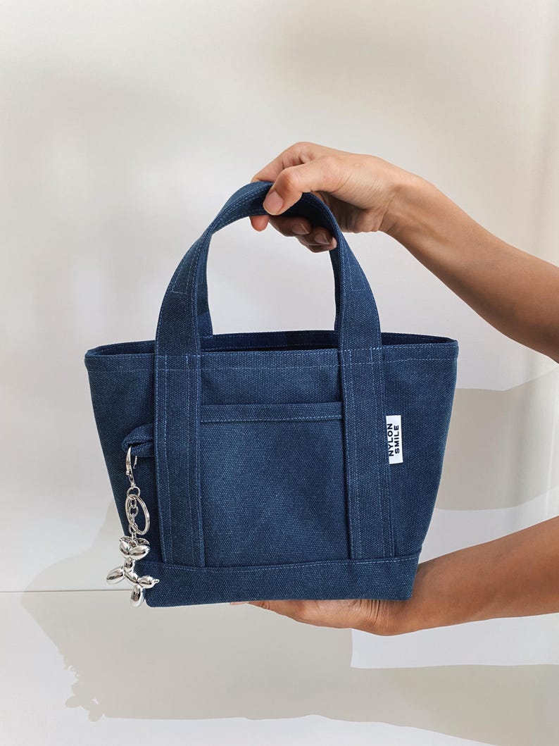 K&ouml;nnte beinhalten: Eine dunkelblaue Tragetasche aus Denim mit zwei stabilen Griffen und einer Au&szlig;entasche auf der Vorderseite. Ein kleines wei&szlig;es rechteckiges Etikett mit dem Text NYLON SMILE ist an der Seite aufgen&auml;ht. Ein silberner Luftballonhund-Anh&auml;nger h&auml;ngt an einem Ring.