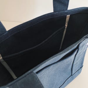 May include: A dark blue canvas tote bag featuring an open top design with two sturdy fabric handles. The interior is lined with matching dark blue fabric and includes a flat side pocket for organization, showcasing a clean and functional structure.