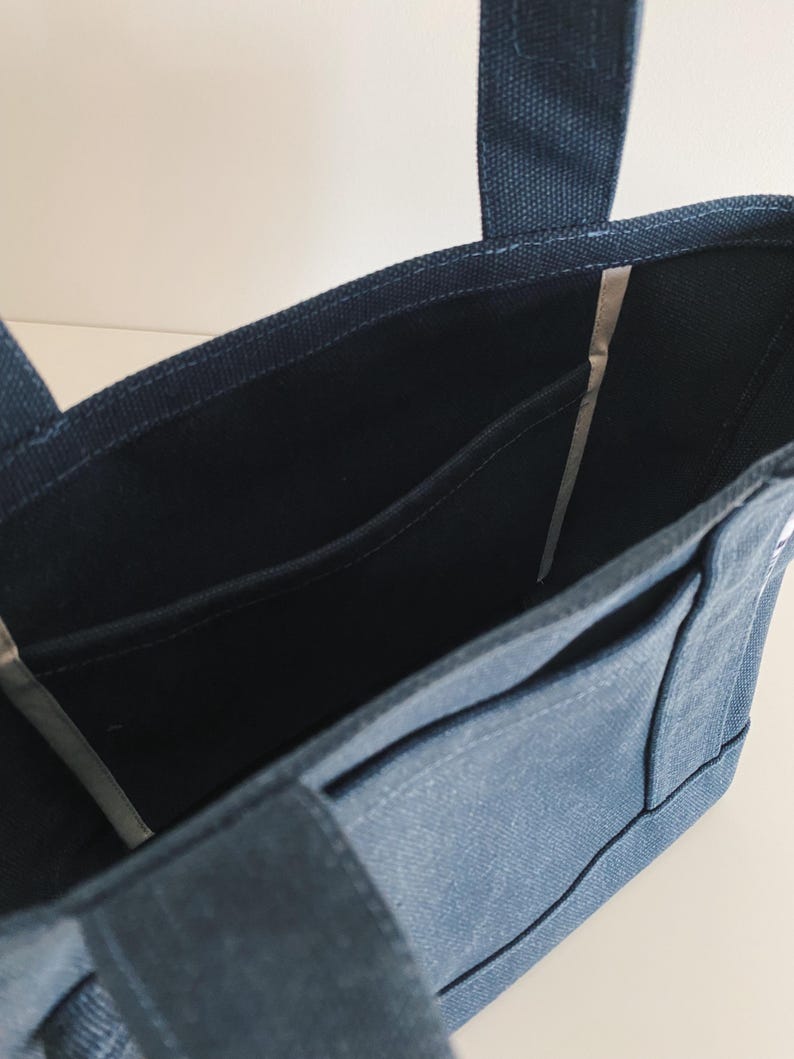 May include: A dark blue canvas tote bag featuring an open top design with two sturdy fabric handles. The interior is lined with matching dark blue fabric and includes a flat side pocket for organization, showcasing a clean and functional structure.