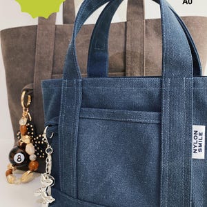 May include: A dark blue canvas tote bag with two handles, a front pocket, and a white label reading NYLON SMILE. A silver balloon dog keychain is attached to the side. Behind it sits a larger brown canvas tote bag with a beaded 8-ball keychain.