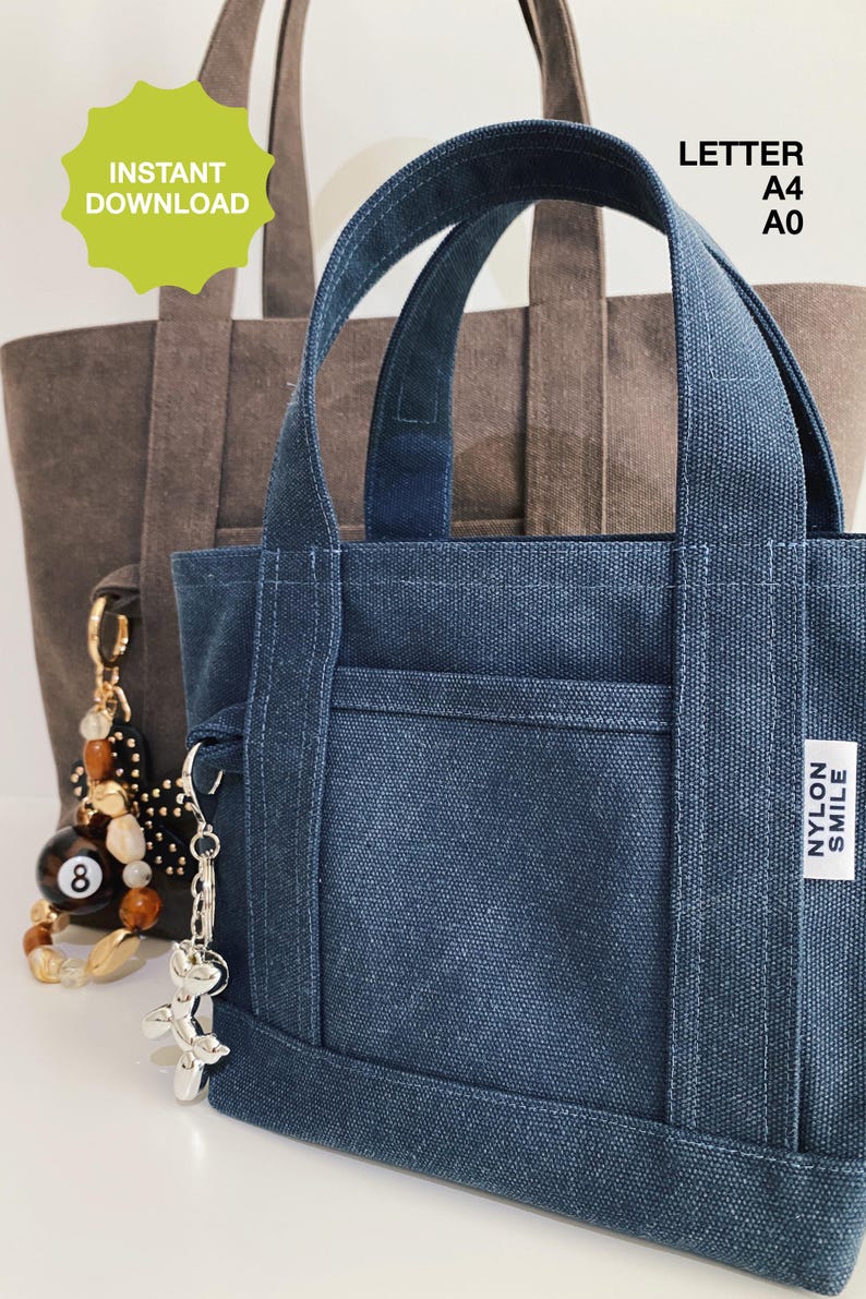 May include: A dark blue canvas tote bag with two handles, a front pocket, and a white label reading NYLON SMILE. A silver balloon dog keychain is attached to the side. Behind it sits a larger brown canvas tote bag with a beaded 8-ball keychain.