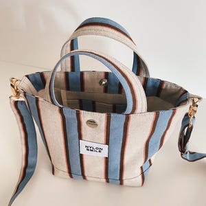 May include: A small tote bag featuring vertical stripes in light blue, brown, and off-white. The bag has two top handles, a detachable shoulder strap with gold-tone hardware, and a white rectangular patch with the text NYLON SMILE.