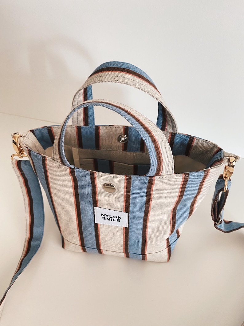 May include: A small tote bag featuring vertical stripes in light blue, brown, and off-white. The bag has two top handles, a detachable shoulder strap with gold-tone hardware, and a white rectangular patch with the text NYLON SMILE.