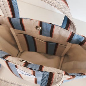 May include: A canvas tote bag featuring vertical stripes in light blue, dark brown, and cream. The bag has two top handles, an open interior with a single slip pocket, and a silver-toned metal snap button closure. A small white rectangular label is visible.