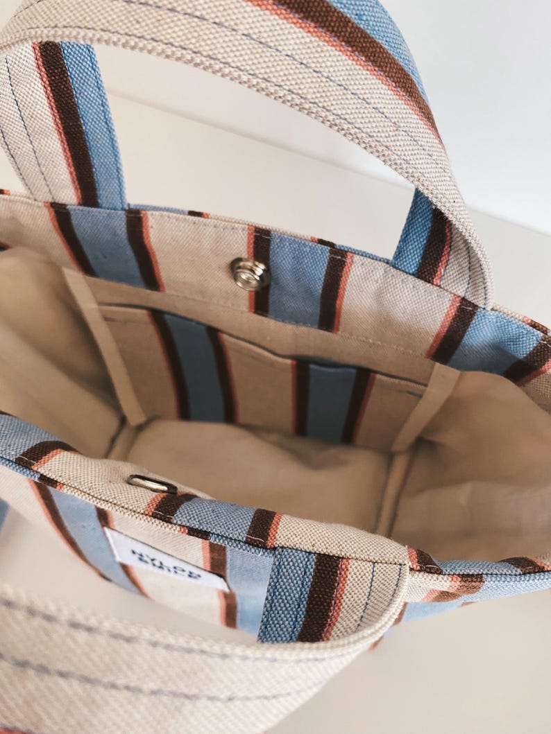May include: A canvas tote bag featuring vertical stripes in light blue, dark brown, and cream. The bag has two top handles, an open interior with a single slip pocket, and a silver-toned metal snap button closure. A small white rectangular label is visible.