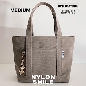 Pode incluir: Uma bolsa tote de tamanho médio na cor taupe com duas alças superiores, um bolso frontal e um pequeno pingente de cachorro de balão dourado na lateral. A bolsa tem uma etiqueta branca com o texto NYLON SMILE e inclui os textos MEDIUM, PDF PATTERN, ENGLISH, ESPAÑOL e NYLON SMILE.