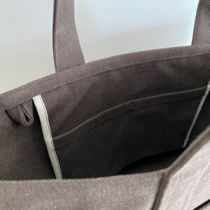 May include: A dark brown canvas tote bag featuring two sturdy shoulder straps, a wide open top, and an interior slip pocket with light-colored trim along the side seams. The fabric has a visible woven texture throughout the exterior and interior.