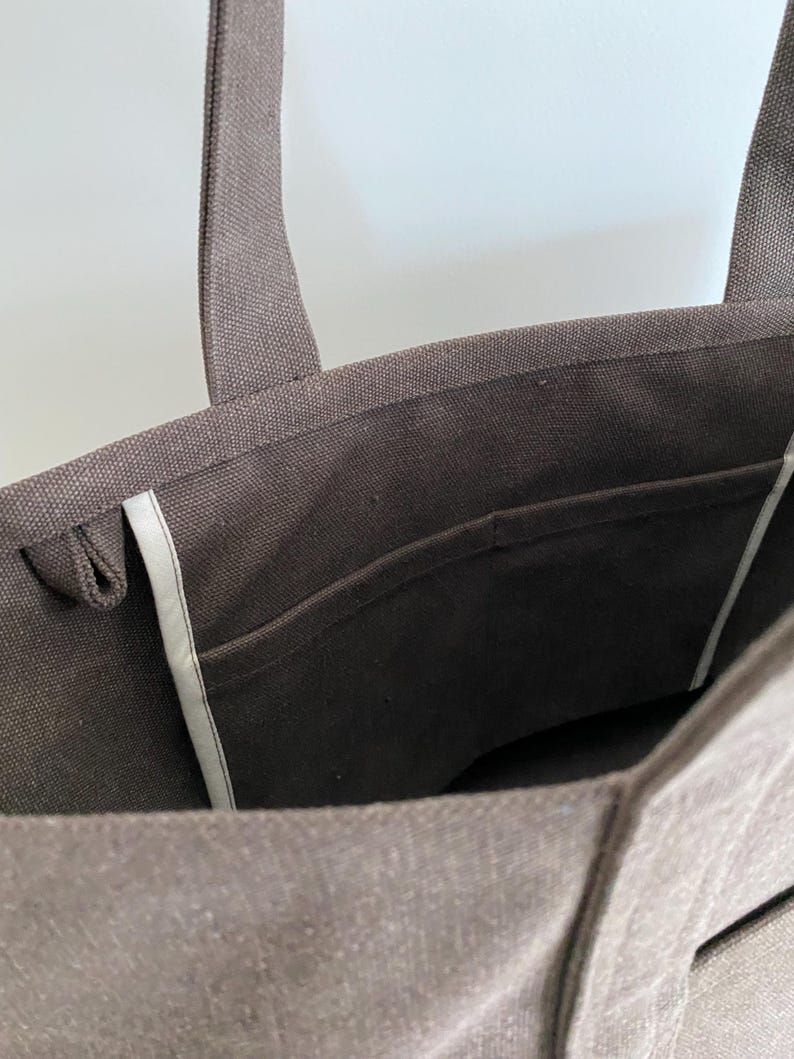 May include: A dark brown canvas tote bag featuring two sturdy shoulder straps, a wide open top, and an interior slip pocket with light-colored trim along the side seams. The fabric has a visible woven texture throughout the exterior and interior.