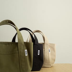 K&ouml;nnte beinhalten: Drei kleine Tragetaschen aus Segeltuch in einer Reihe, in den Farben Olivgr&uuml;n, Schwarz und Beige. Jede Tasche hat zwei stabile Griffe und ein kleines wei&szlig;es rechteckiges Etikett auf der Vorderseite mit dem Wort NUNE in fetten schwarzen Gro&szlig;buchstaben.