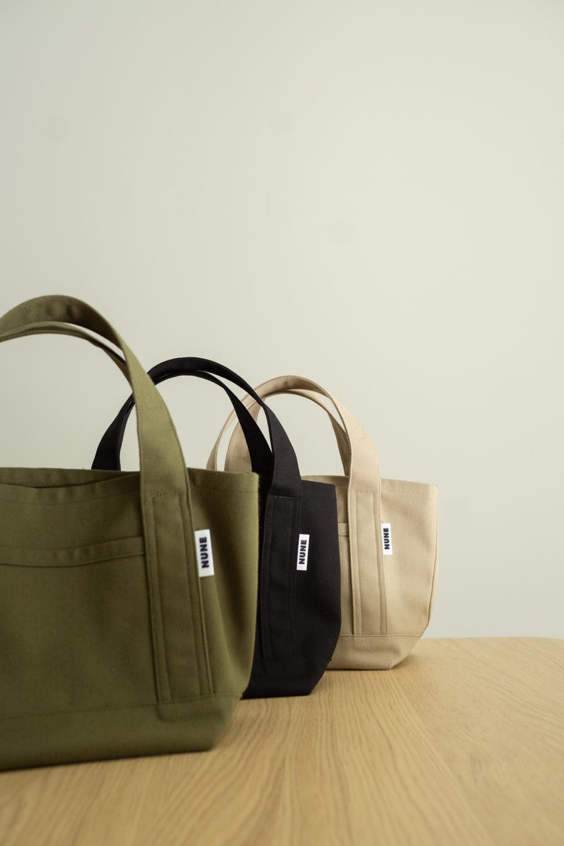 K&ouml;nnte beinhalten: Drei kleine Tragetaschen aus Segeltuch in einer Reihe, in den Farben Olivgr&uuml;n, Schwarz und Beige. Jede Tasche hat zwei stabile Griffe und ein kleines wei&szlig;es rechteckiges Etikett auf der Vorderseite mit dem Wort NUNE in fetten schwarzen Gro&szlig;buchstaben.