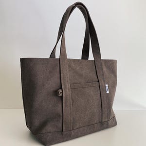 May include: A dark brown canvas tote bag featuring two sturdy shoulder straps, a front exterior pocket, and a small white fabric label on the side with the text "ATELIER SMILE" printed in black. The bag has a simple, structured rectangular design.