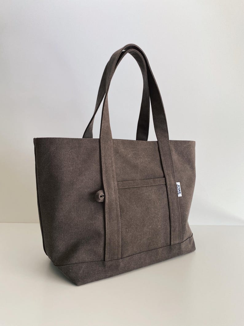 May include: A dark brown canvas tote bag featuring two sturdy shoulder straps, a front exterior pocket, and a small white fabric label on the side with the text "ATELIER SMILE" printed in black. The bag has a simple, structured rectangular design.