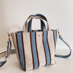 May include: A structured canvas tote bag featuring vertical stripes in light blue, dark brown, and cream. The bag includes two top handles, a central silver-tone snap button closure, and a detachable, adjustable shoulder strap with gold-tone hardware.