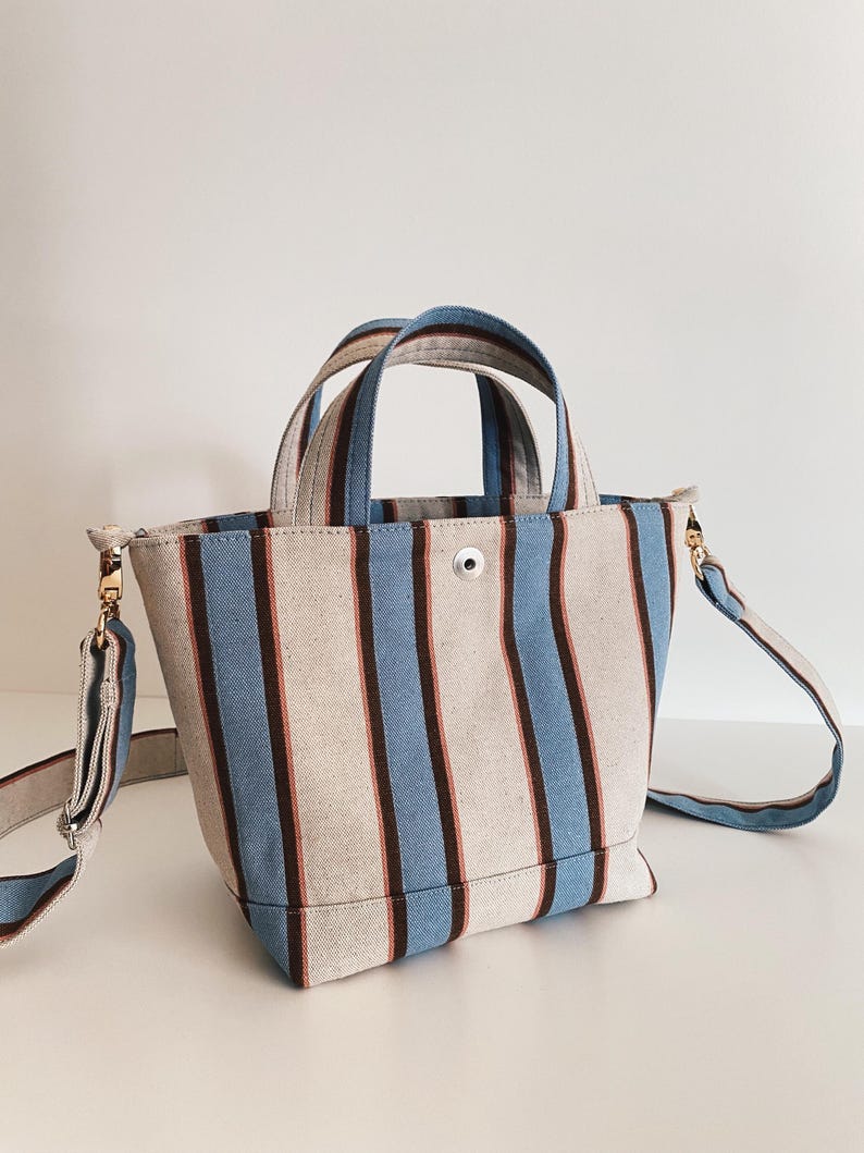 May include: A structured canvas tote bag featuring vertical stripes in light blue, dark brown, and cream. The bag includes two top handles, a central silver-tone snap button closure, and a detachable, adjustable shoulder strap with gold-tone hardware.