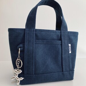May include: A dark blue canvas tote bag with two top handles and a front slip pocket. A small white rectangular tag on the side reads NYLON SMILE. A silver-colored metallic balloon dog keychain is attached to the side of the bag by a metal ring.